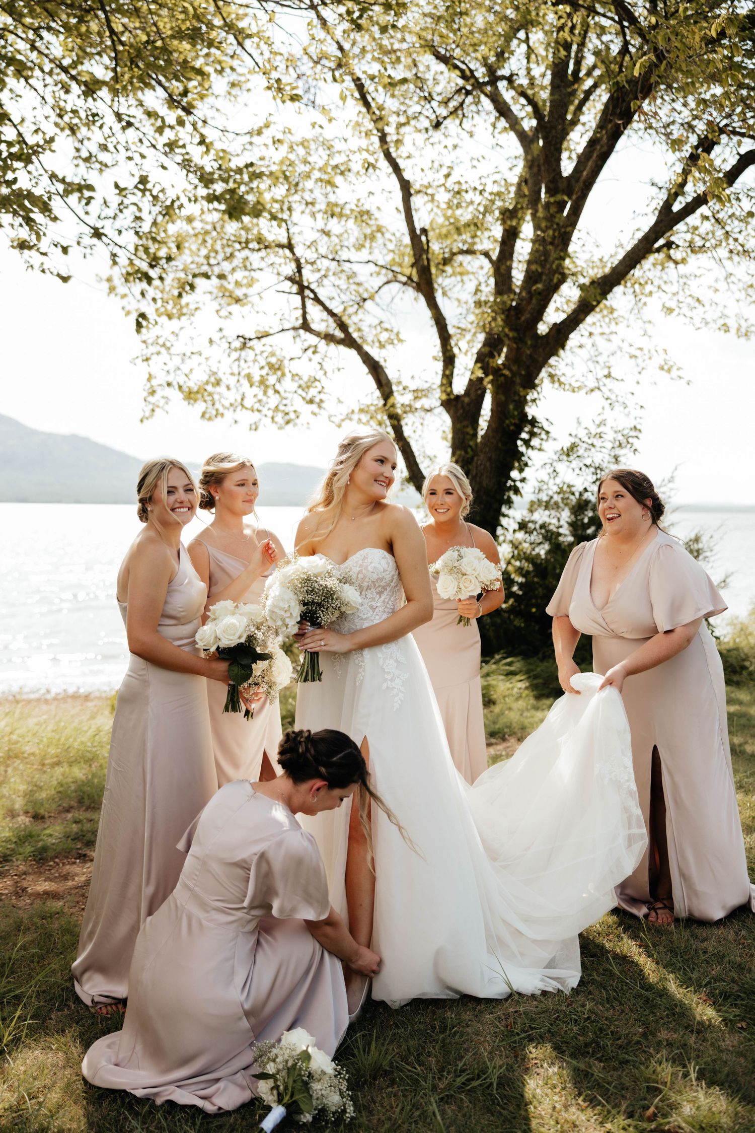 Bride in strapless lace gown poses with five bridesmaids in blush dresses near a lakeside tree.