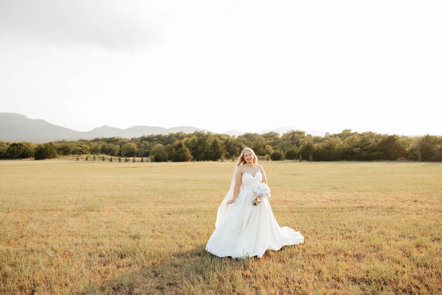 Radiant bride in flowing white gown holding bouquet stands alone in golden open field with mountain backdrop.