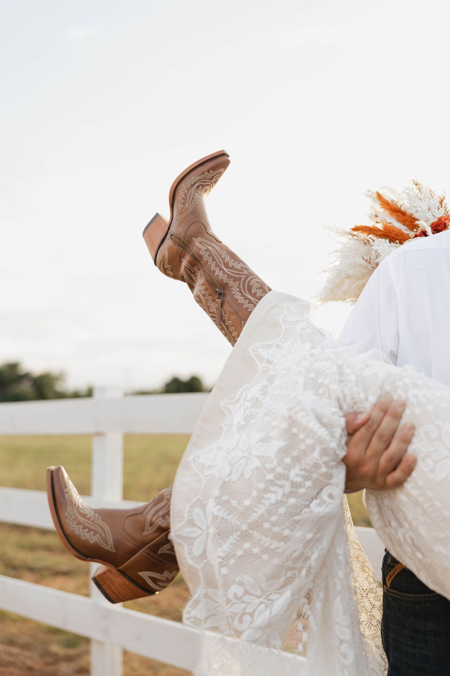Close-up of bride's tan cowboy boots and lace wedding dress held by groom near white farm fence.