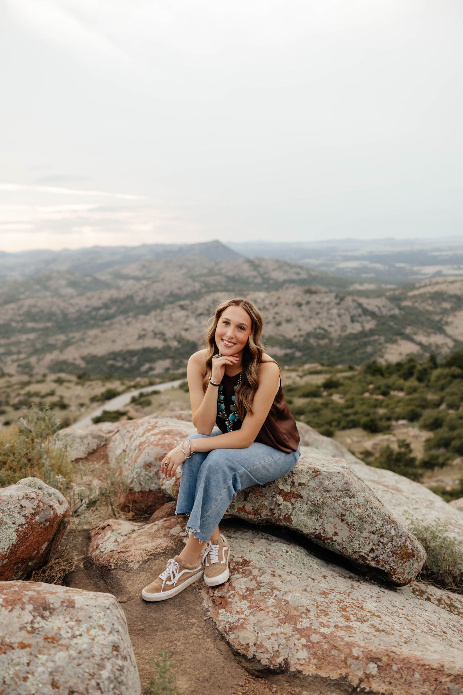 Young woman sitting on rocks with scenic mountain landscape backdrop, smiling in jeans and dark floral top.