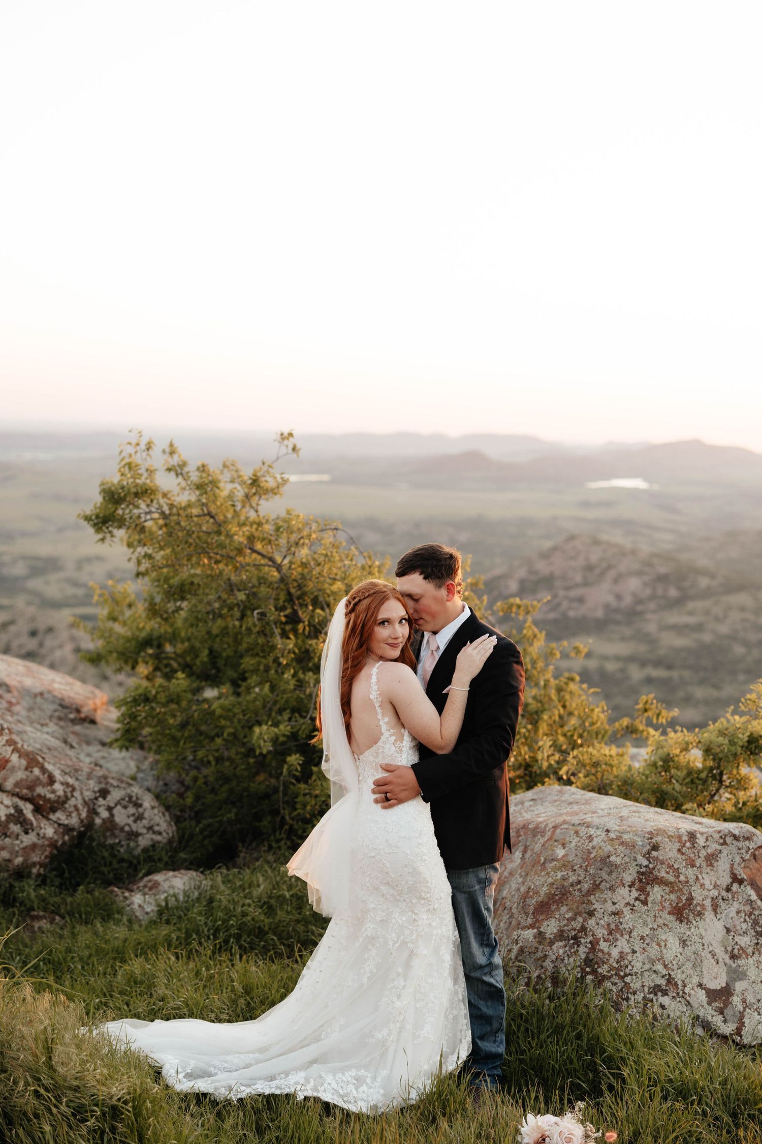 Bride and groom embrace on a scenic mountain overlook at sunset in wedding attire.