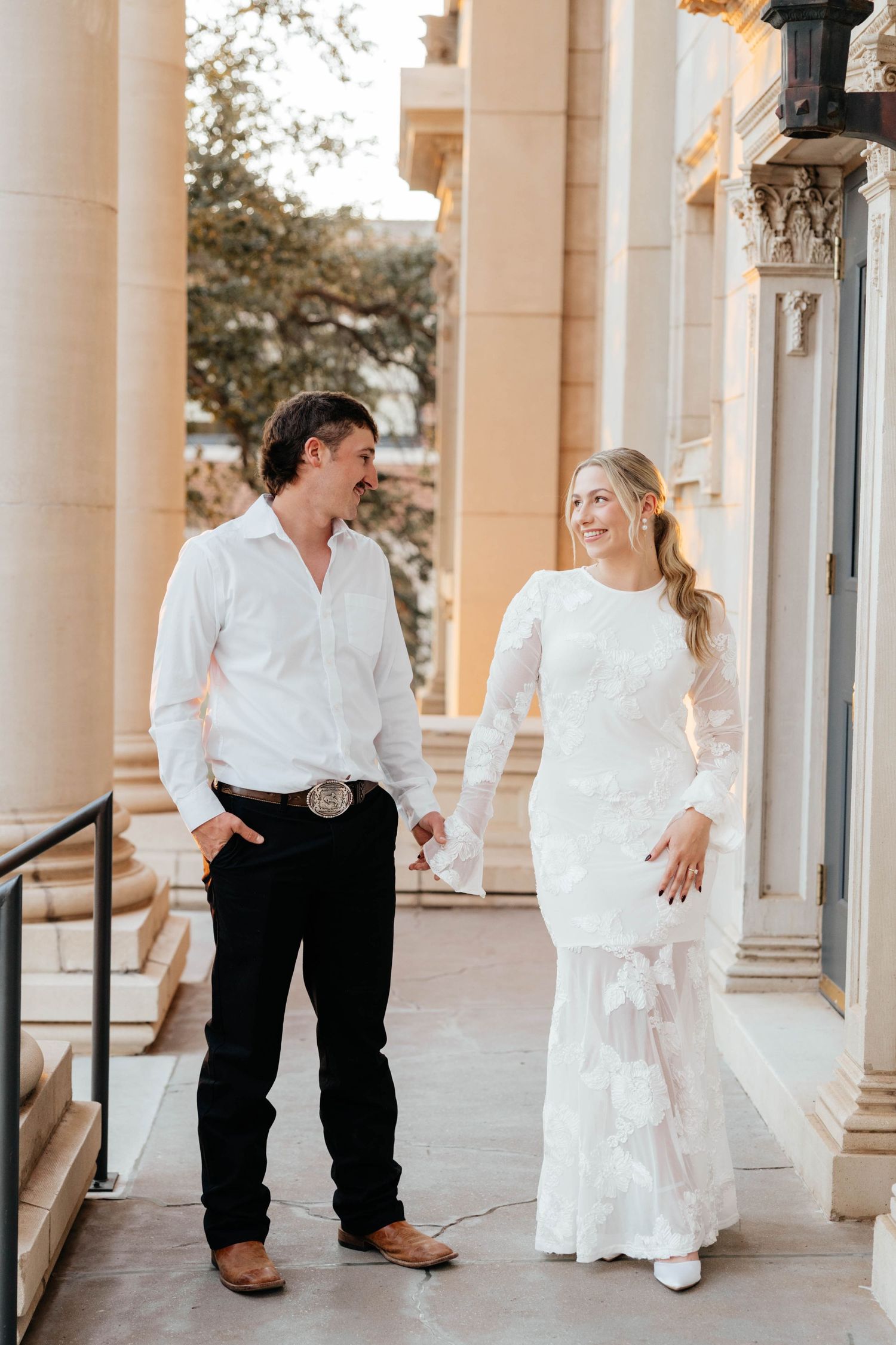 Bride in long-sleeve lace gown and groom in white shirt with belt buckle walk between stone columns.