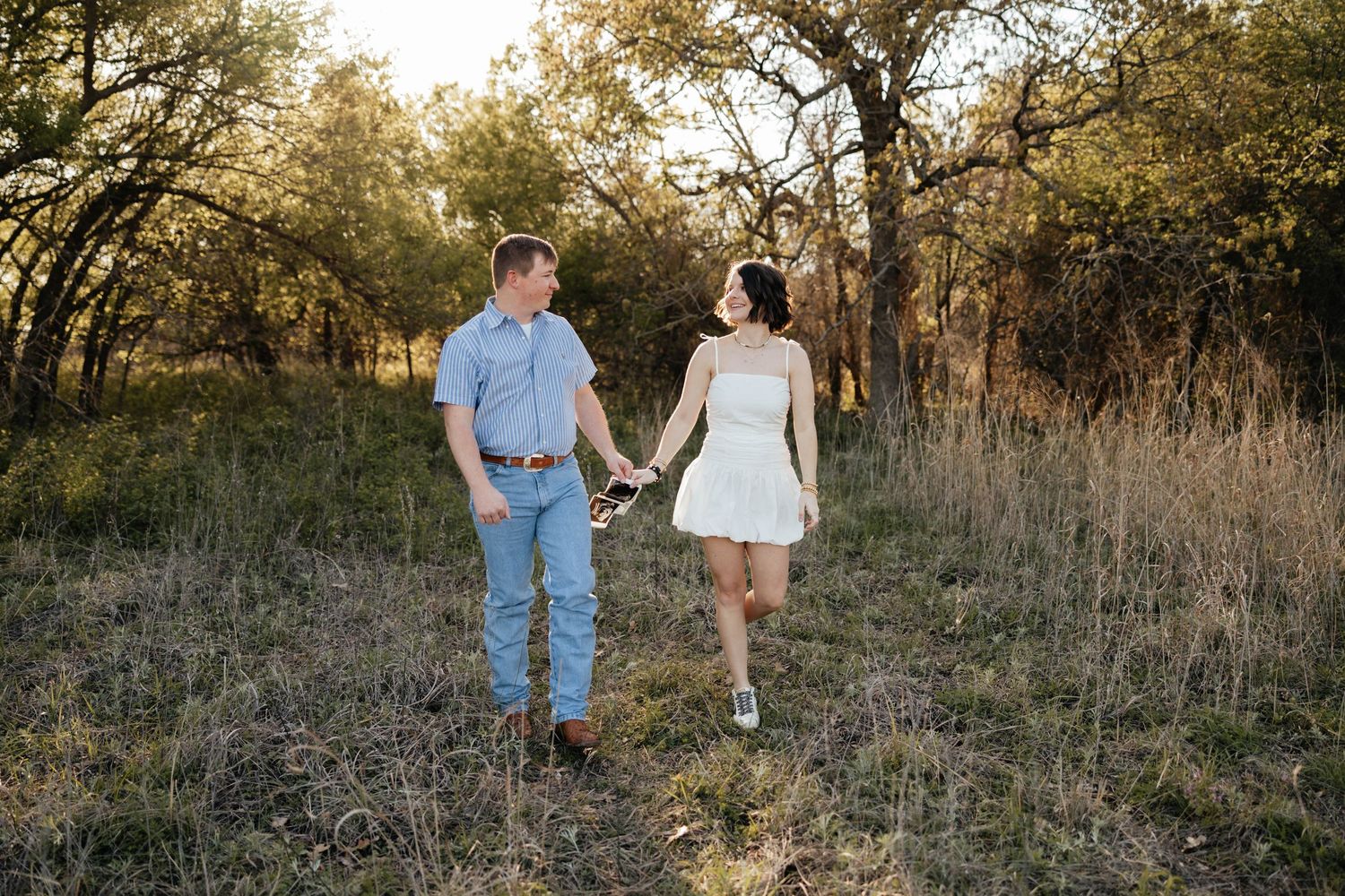 Couple holding hands and walking through a sunlit wooded meadow during golden hour engagement session.