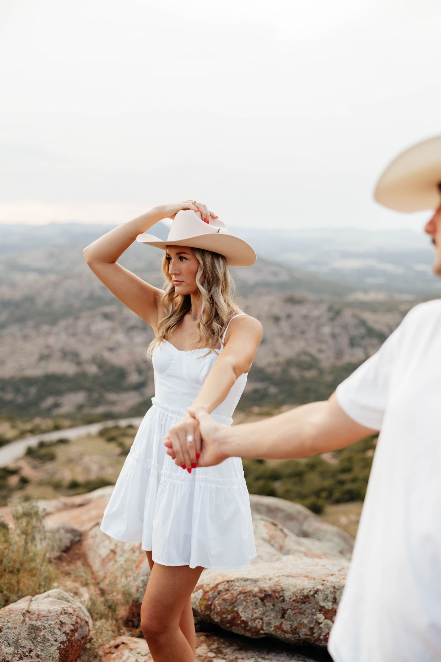 Blonde woman in white sundress and tan cowboy hat holding hands with man on scenic mountain overlook.