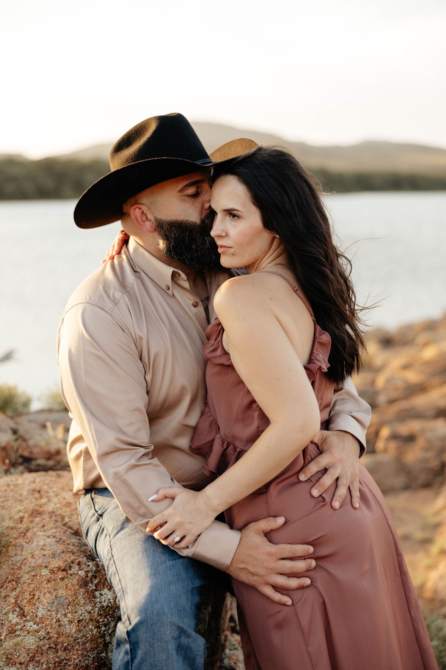Couple shares intimate embrace on rocky lakeside, man in black cowboy hat kissing woman in mauve dress.