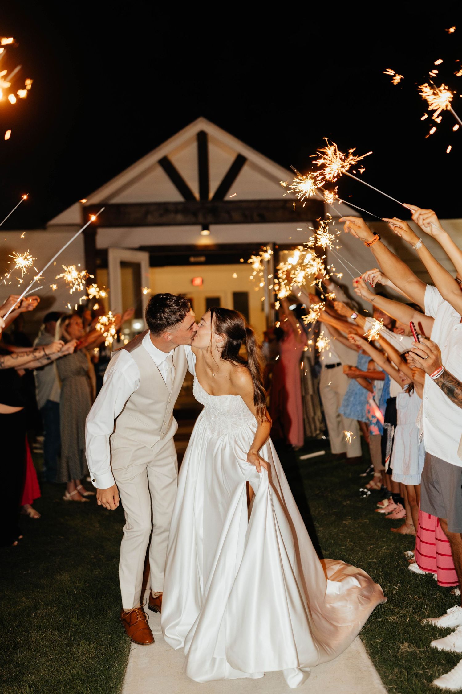 Newlyweds share a kiss during a romantic sparkler send-off exit at night outside a white wedding venue.