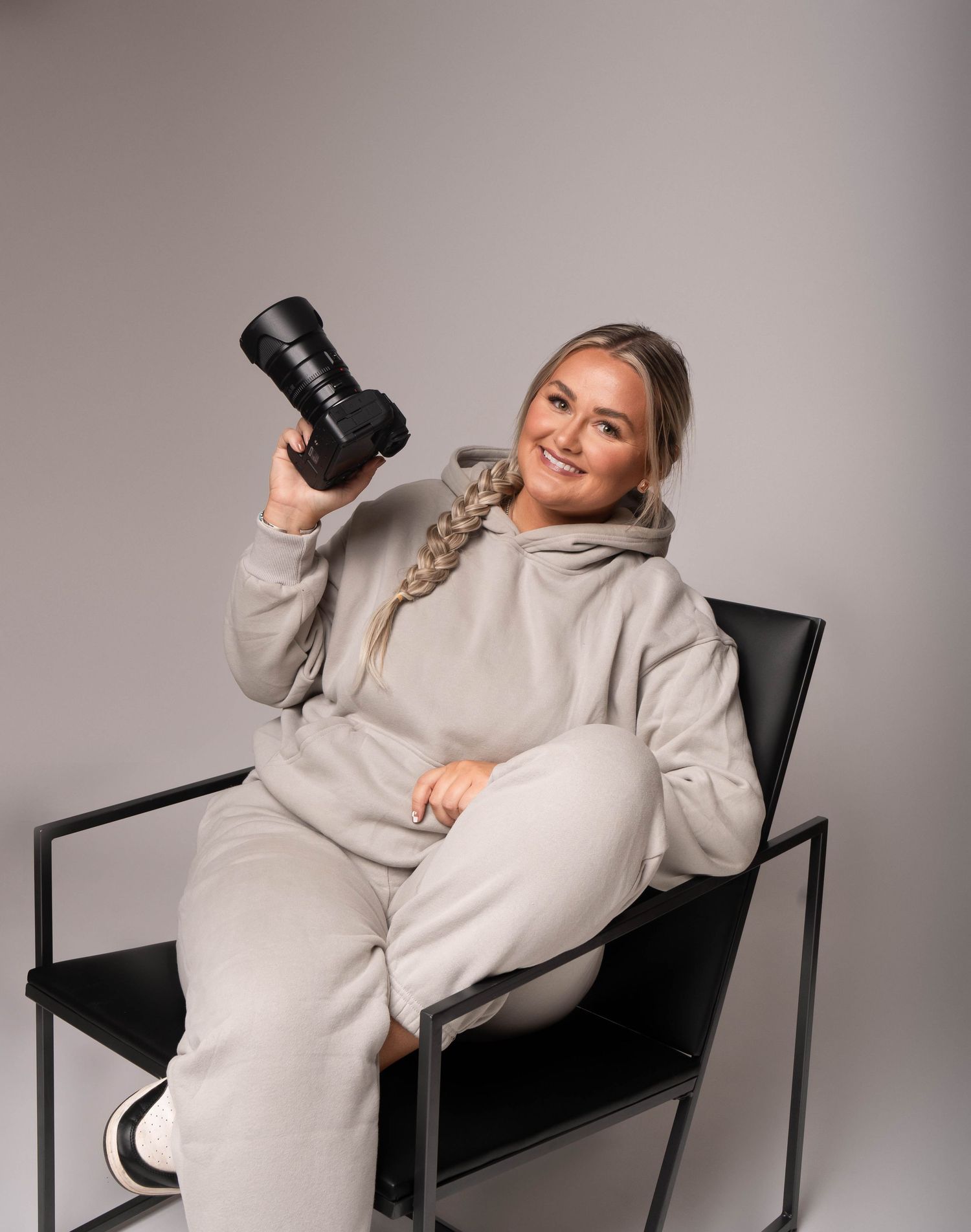 Smiling female photographer in beige hoodie poses with professional camera while seated in a black chair.