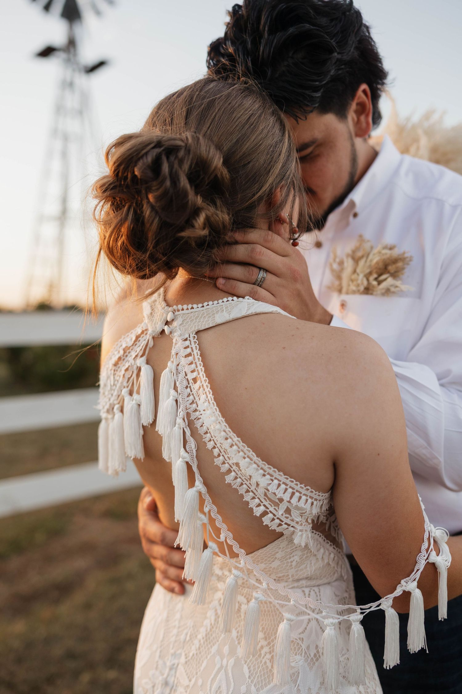 Groom kisses bride's head showing open-back boho lace wedding dress with fringe near windmill.
