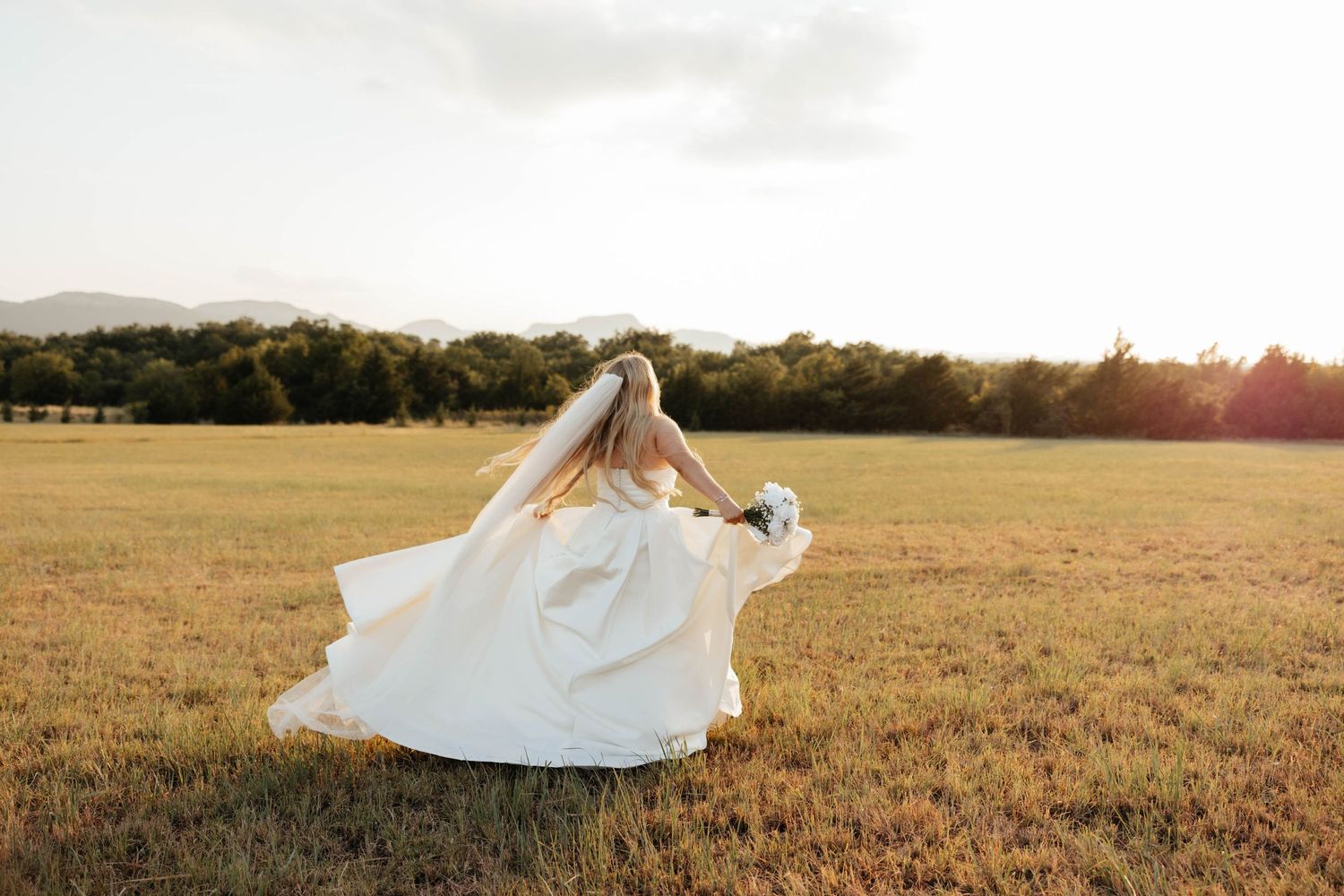 Bride in a flowing white gown and veil twirls in a golden sunlit open field at sunset.