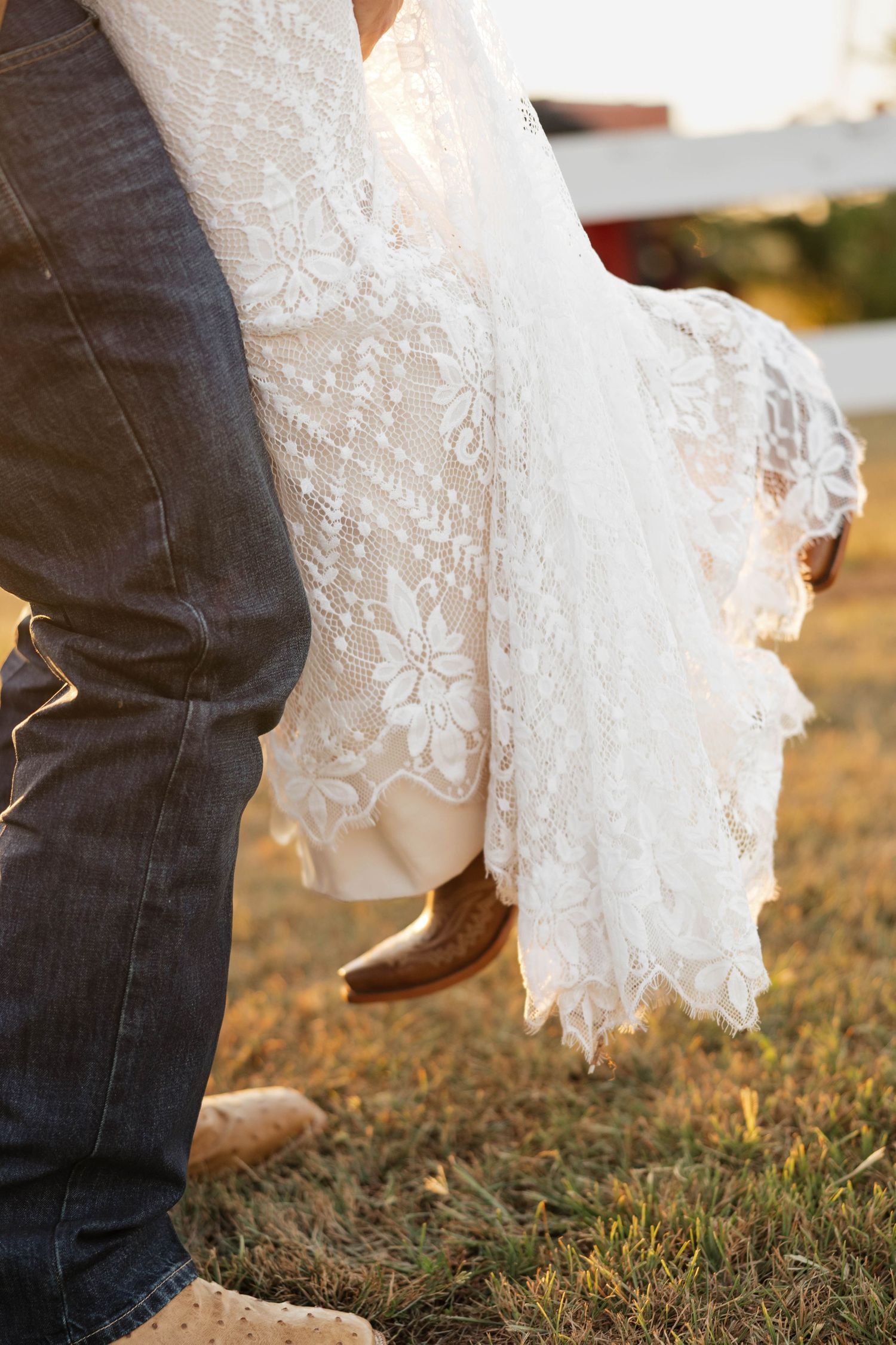 Groom in jeans carries bride in floral lace wedding dress and cowboy boots at golden hour.