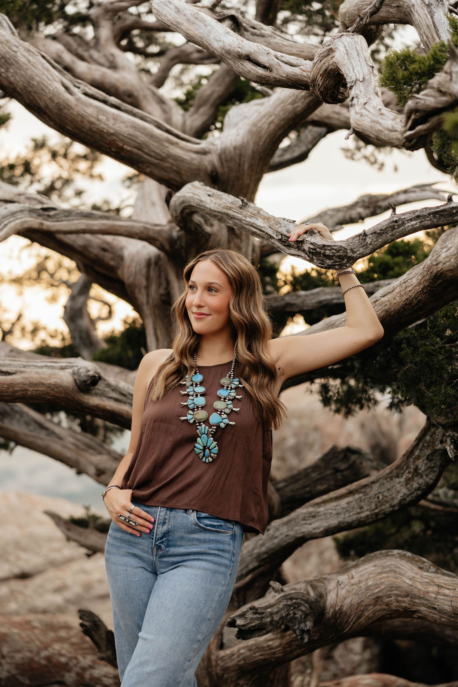 Woman leaning against twisted tree branches wearing turquoise squash blossom necklace and brown tank top.
