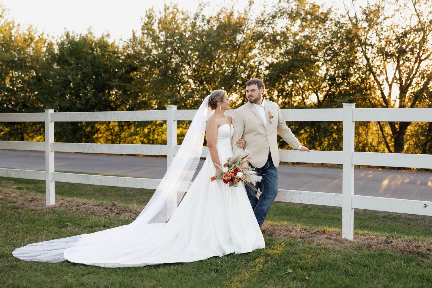 Bride and groom share a tender moment by a white fence surrounded by golden autumn trees.