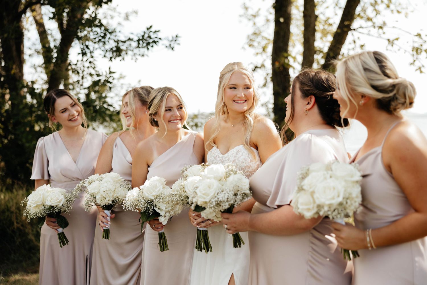 Bridesmaids in taupe dresses holding white floral bouquets smile together outdoors at a wedding.