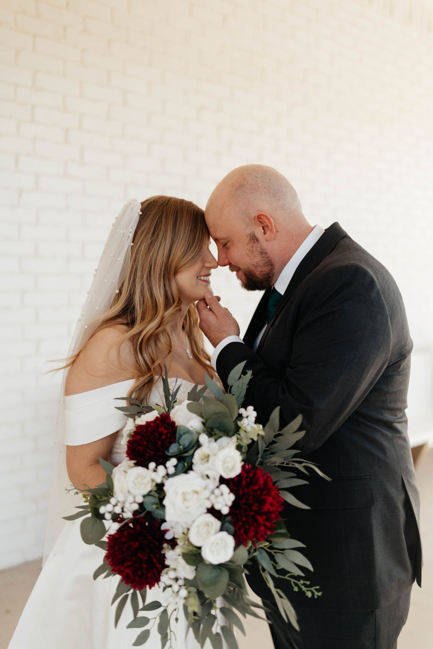 Bride and groom touch foreheads tenderly holding a red and white floral bouquet indoors.