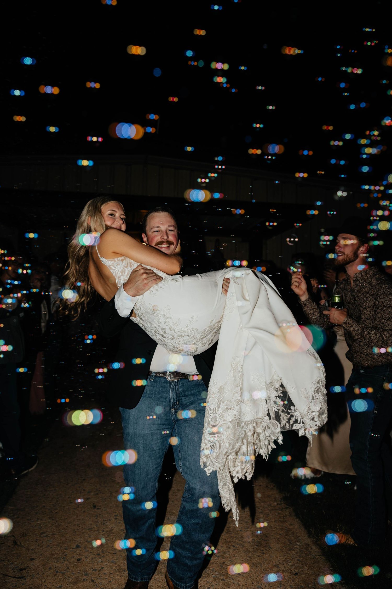 Groom carrying bride through colorful bubble send-off at outdoor wedding reception at night.