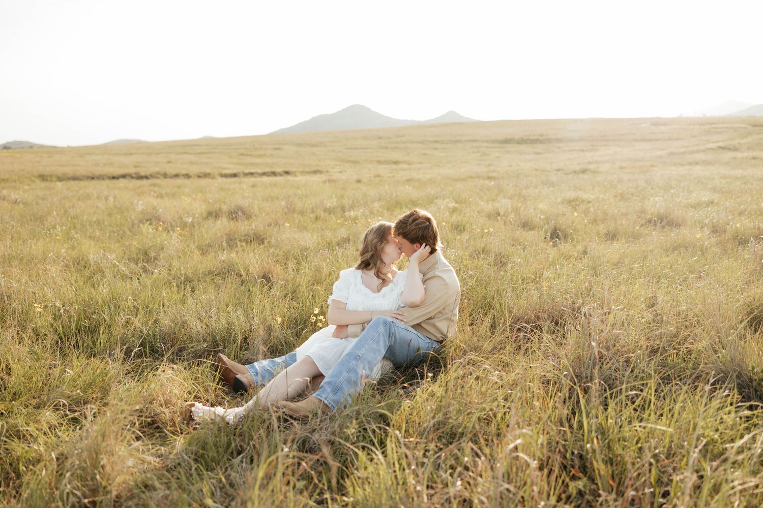 Couple sitting together in a golden grass field with mountains in the background at sunset.