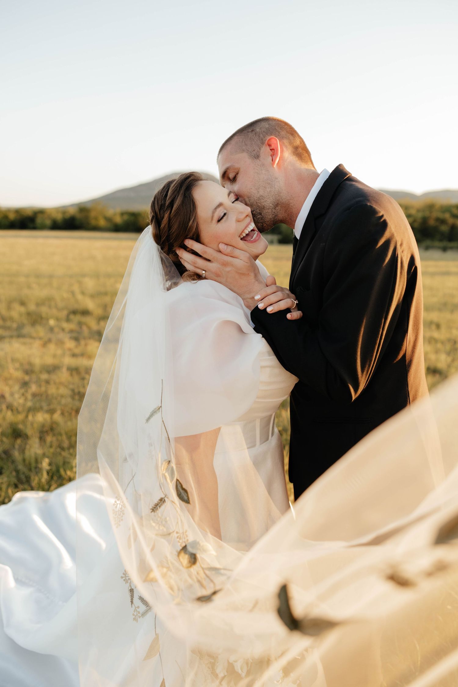 Groom tenderly kissing brides cheek while holding her face in sunlit open field at sunset.