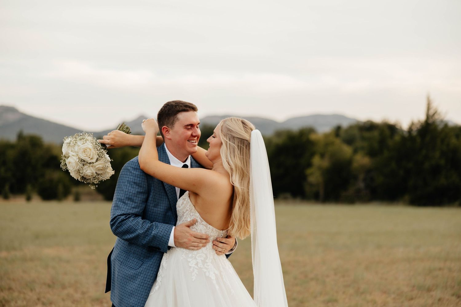 Bride and groom embracing in a field with mountain backdrop, bride holding white bouquet in wedding attire.