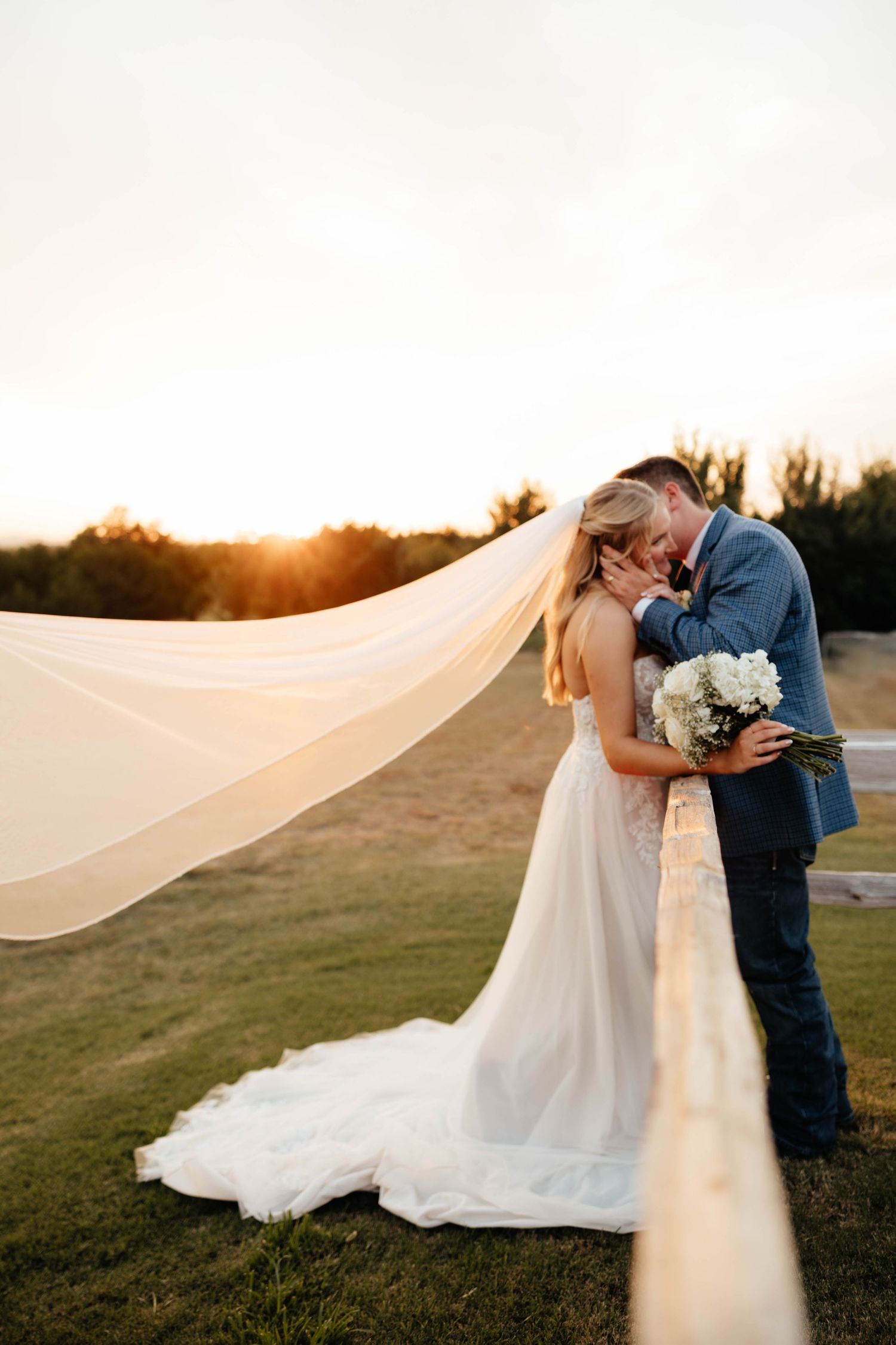 Bride and groom share a sunset kiss outdoors, with her veil flowing dramatically in the wind.
