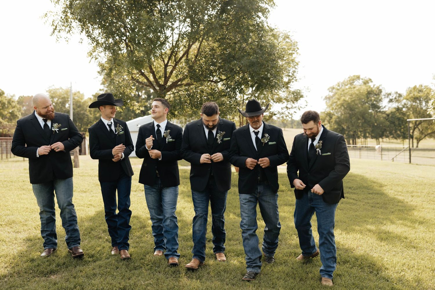 Groomsmen in black blazers and jeans walk together across a green lawn at an outdoor wedding.