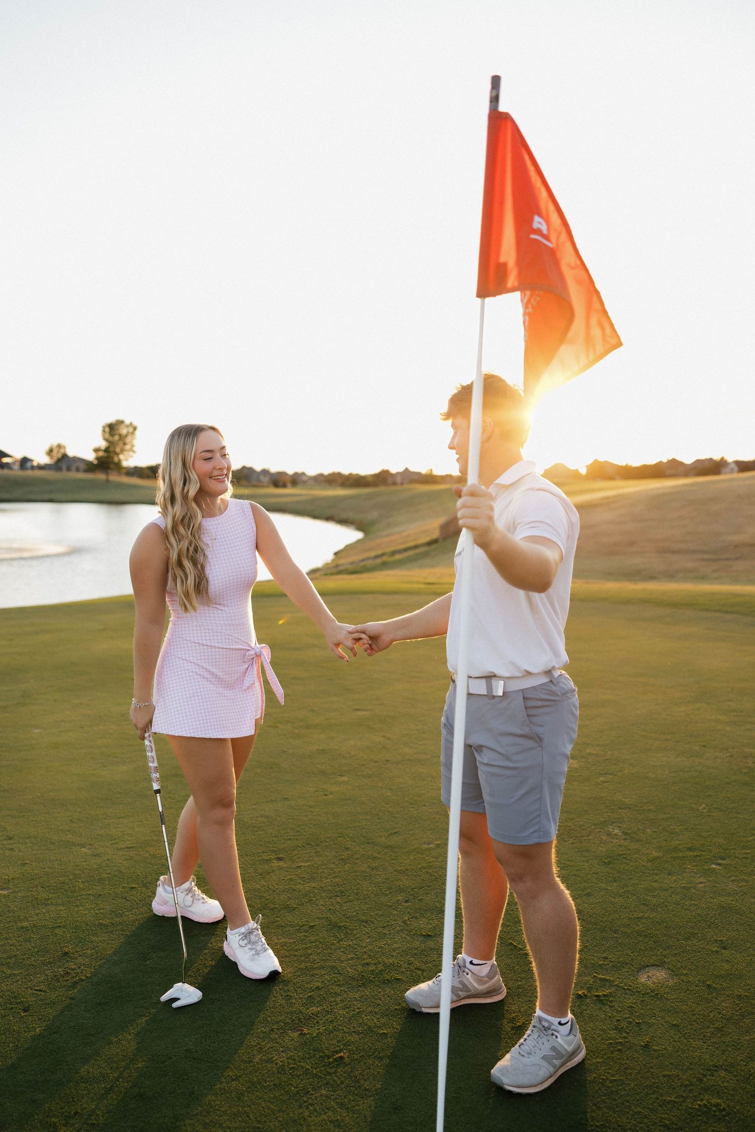 Engaged couple holding hands on golf course at sunset with red flag and water in background.