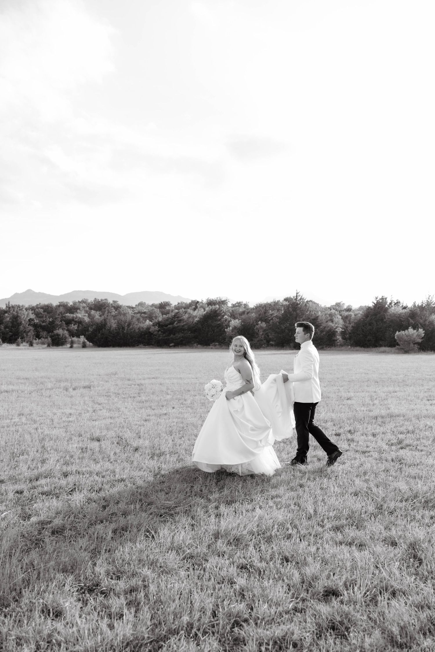 Black and white photo of bride and groom walking through an open field with mountain backdrop.
