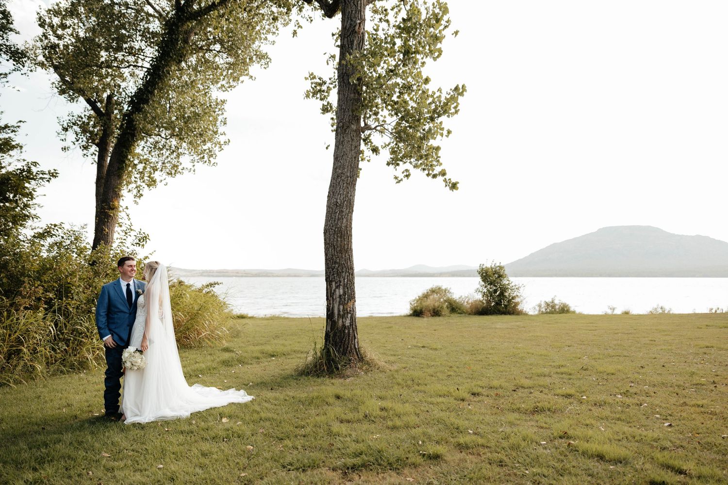 Bride and groom stand together near tall trees with a serene lake and mountain backdrop.