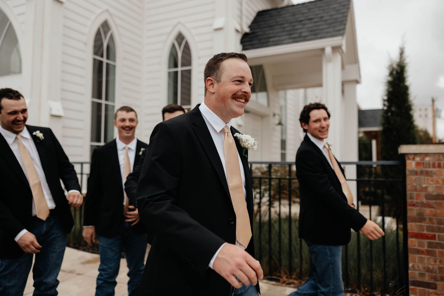 Groom and groomsmen in black suits with tan ties laughing outside a white chapel on a wedding day.