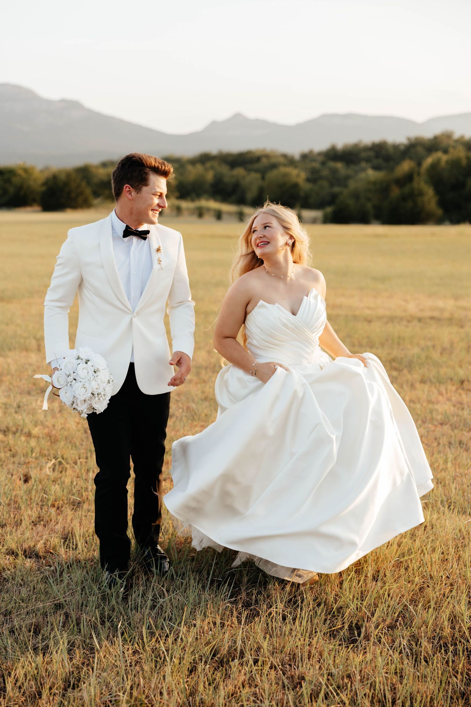 Laughing bride and groom walk through a golden field at sunset, holding hands joyfully.