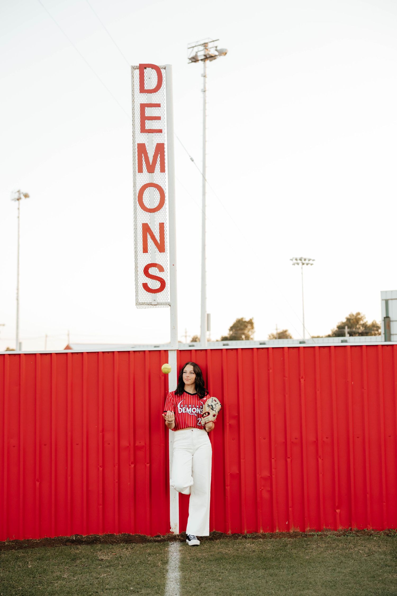 Female softball player in red Demons jersey poses by vertical team sign at red fence.