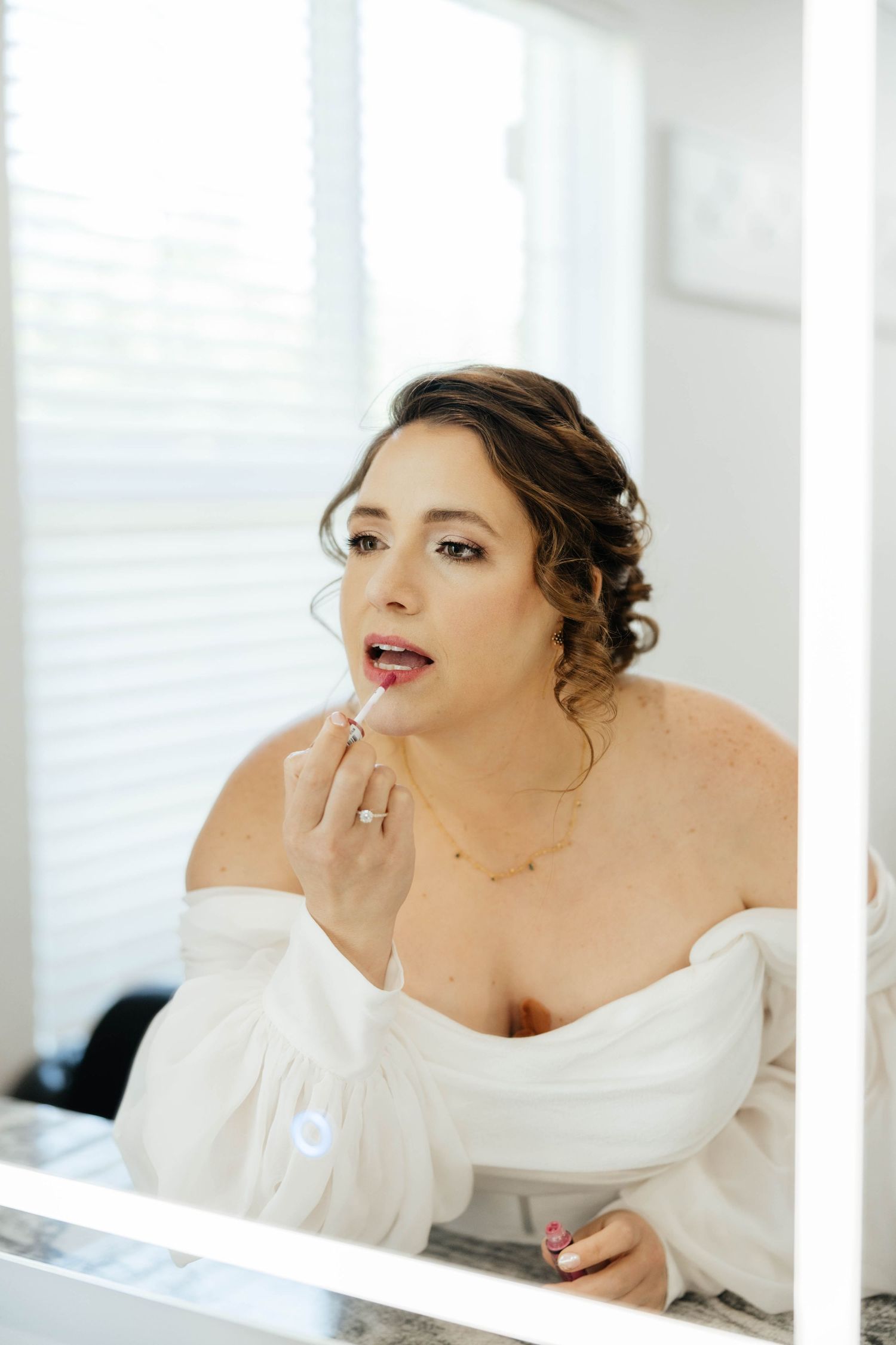 Bride applying lipstick while looking in mirror wearing off-shoulder white robe before wedding.