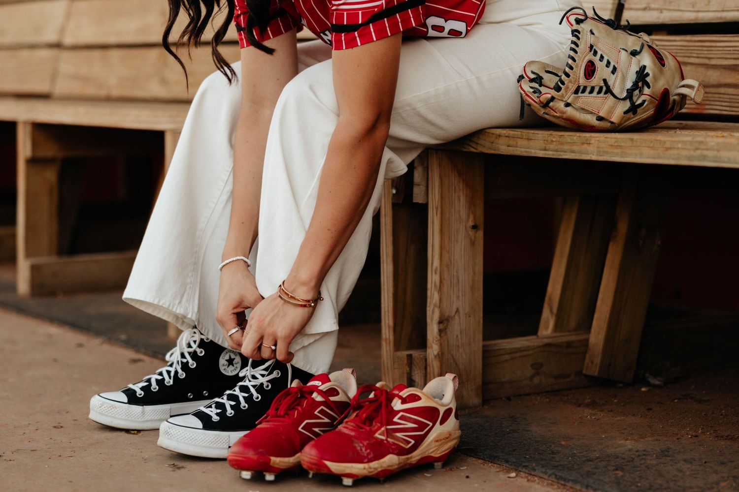 Close-up of softball player tying black Converse sneakers beside red cleats on wooden bench.