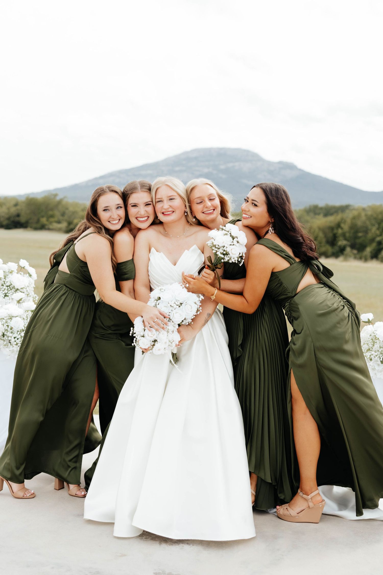 Bride in white gown surrounded by four bridesmaids in olive green dresses outdoors.