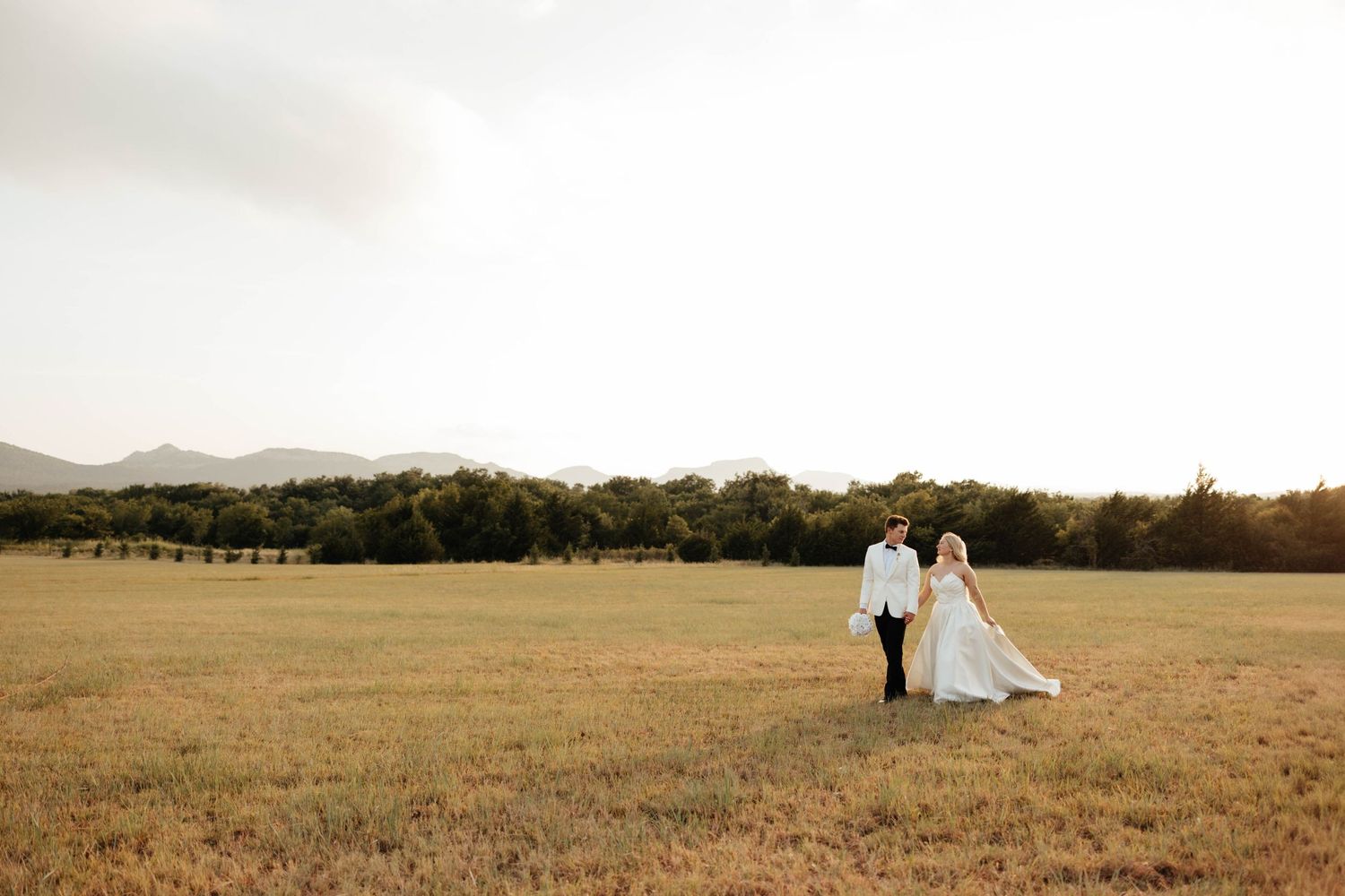 Bride and groom walk hand in hand through a golden field with mountains in the background.