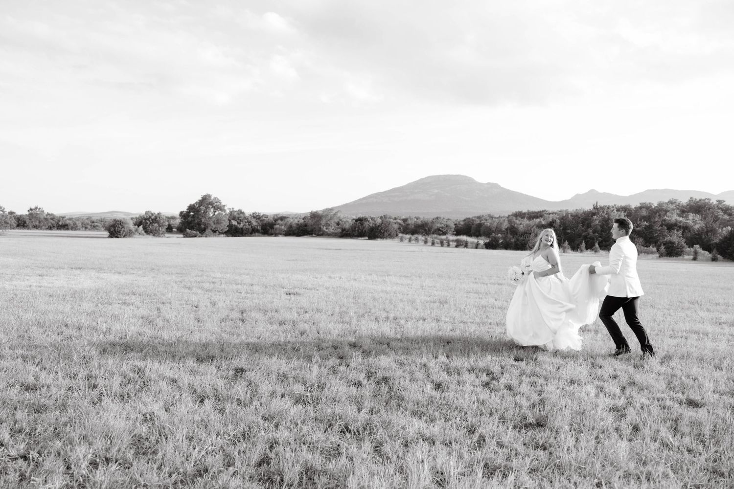 Black and white photo of bride and groom walking hand in hand across a vast open field with mountains.