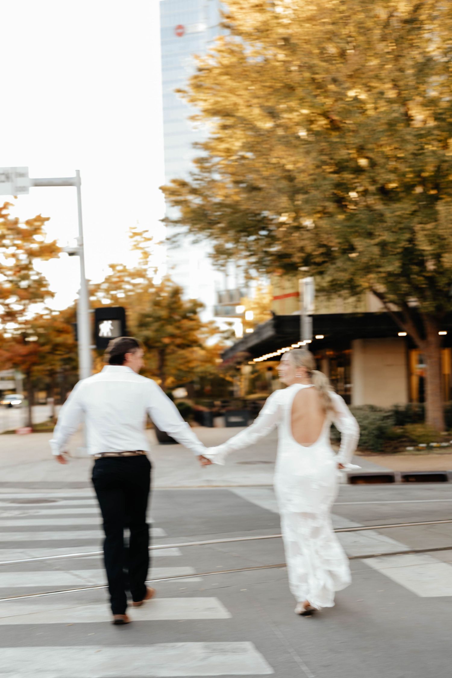 Bride and groom holding hands crossing city street at sunset in motion-blurred wedding photo.