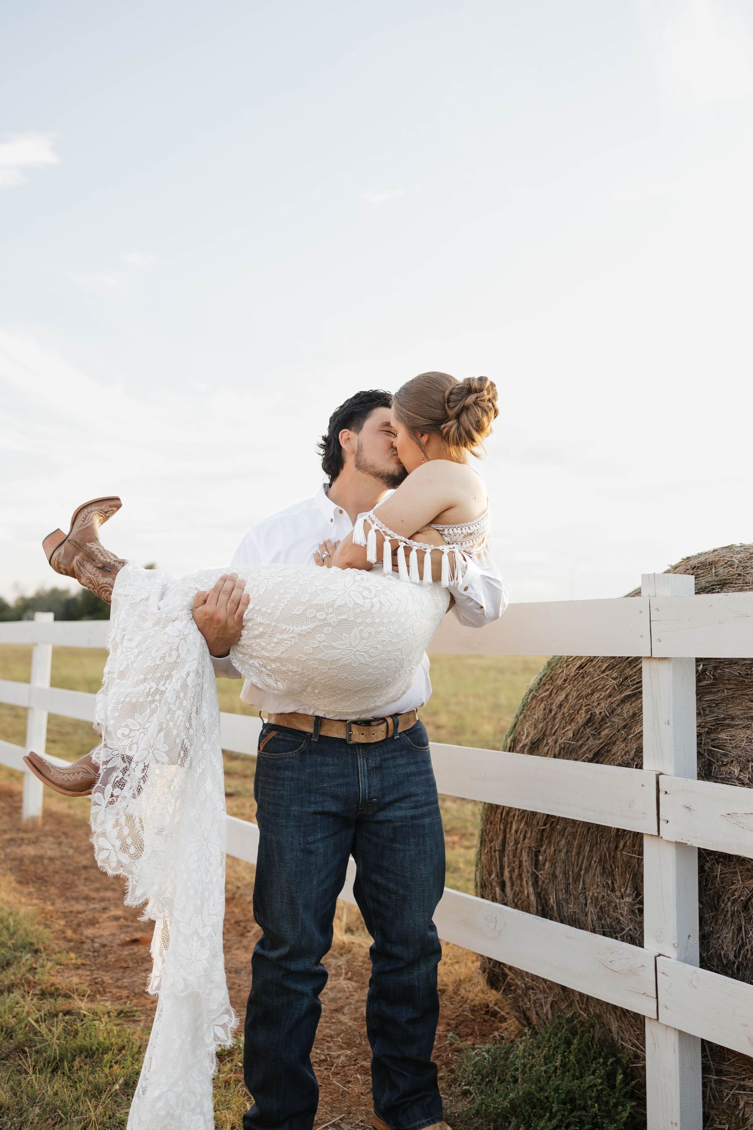 Groom carries bride in lace dress and cowboy boots while kissing near white farm fence with hay bale.