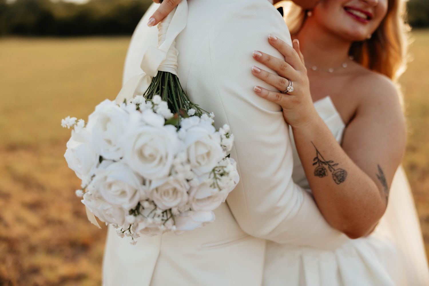Bride in strapless gown with tattoo holds white rose bouquet while embracing groom in sunlit field.