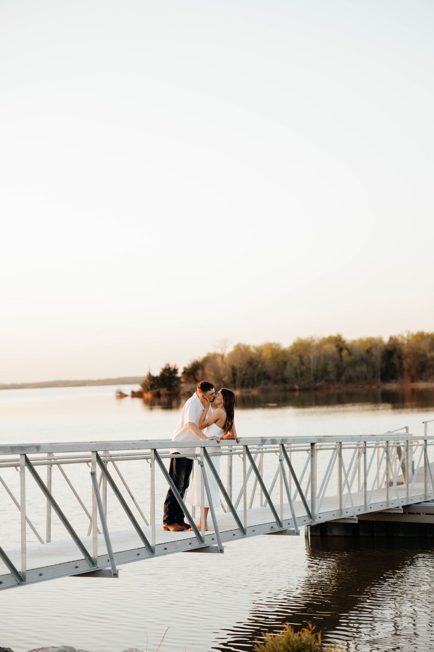 Couple shares a romantic kiss on a white dock overlooking a calm lake at sunset.