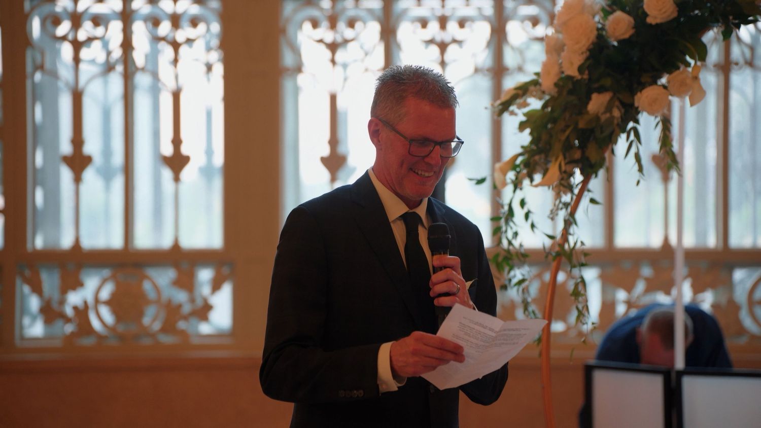 Man in black suit reading speech at elegant wedding reception with floral arch decoration.
