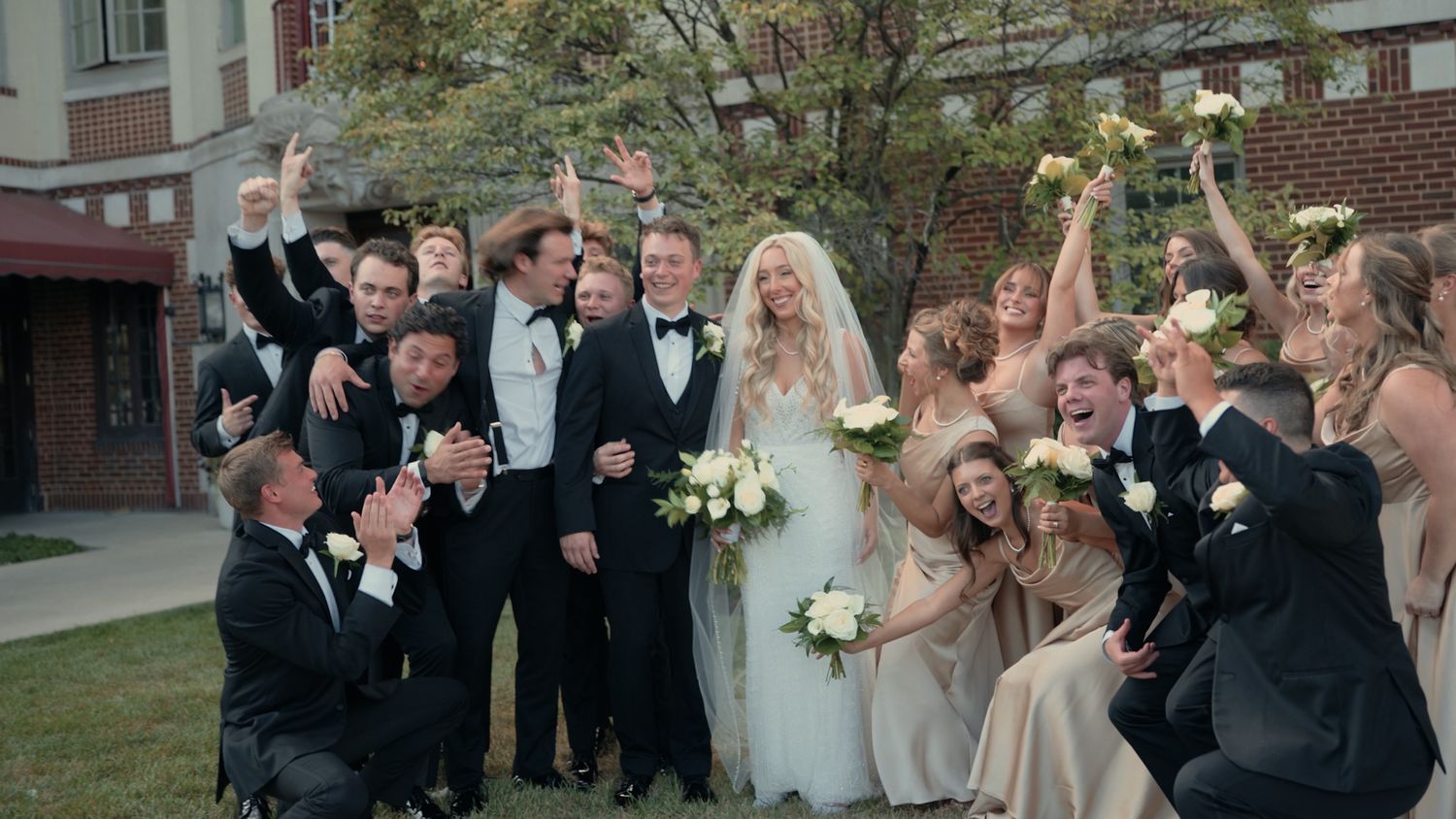A joyful wedding party celebrates outdoors, with bride in white, groom in tuxedo, and cheering bridesmaids and groomsmen.