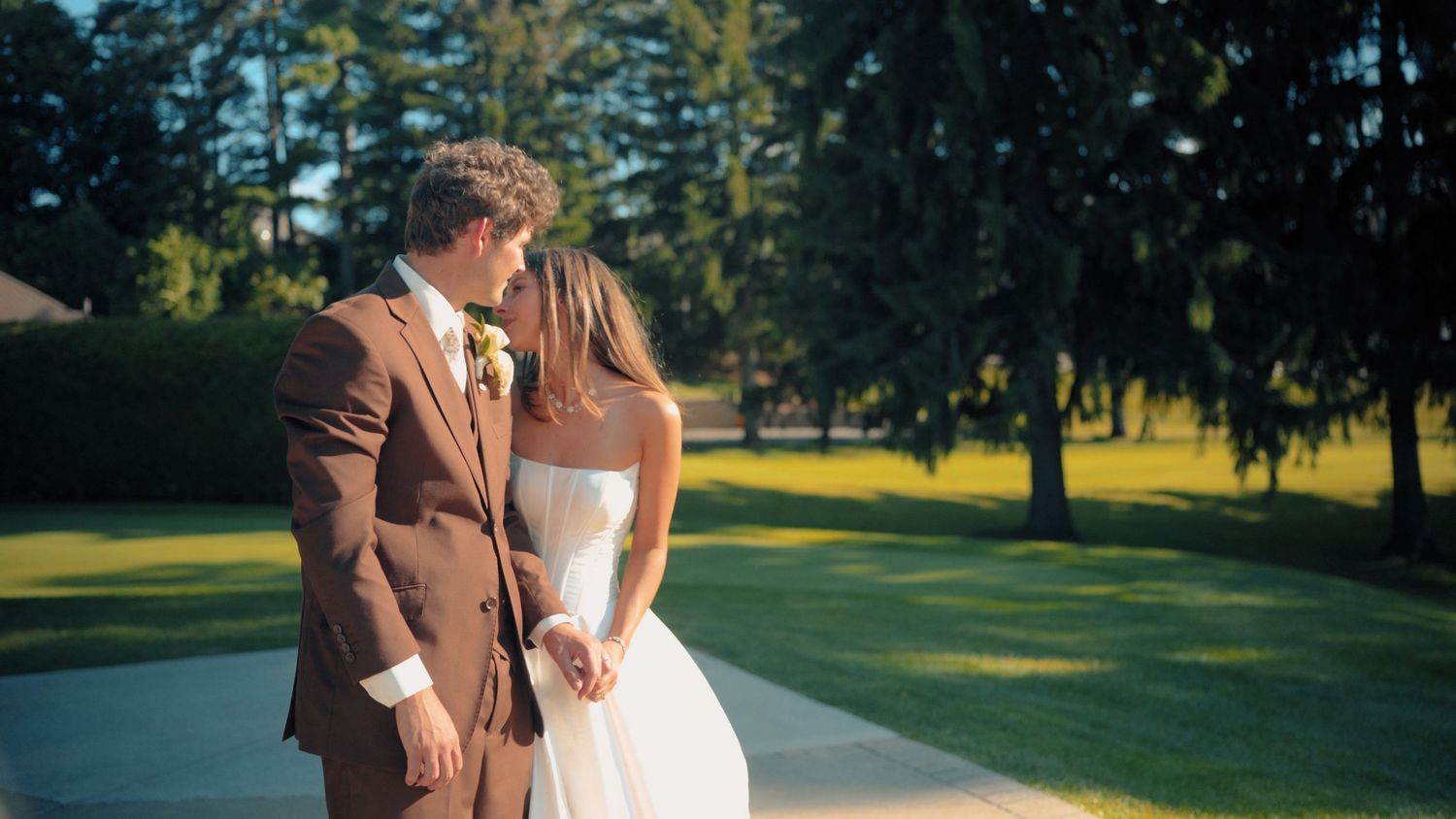 A bride and groom share a romantic moment outdoors on their wedding day, surrounded by lush green trees and manicured lawn.