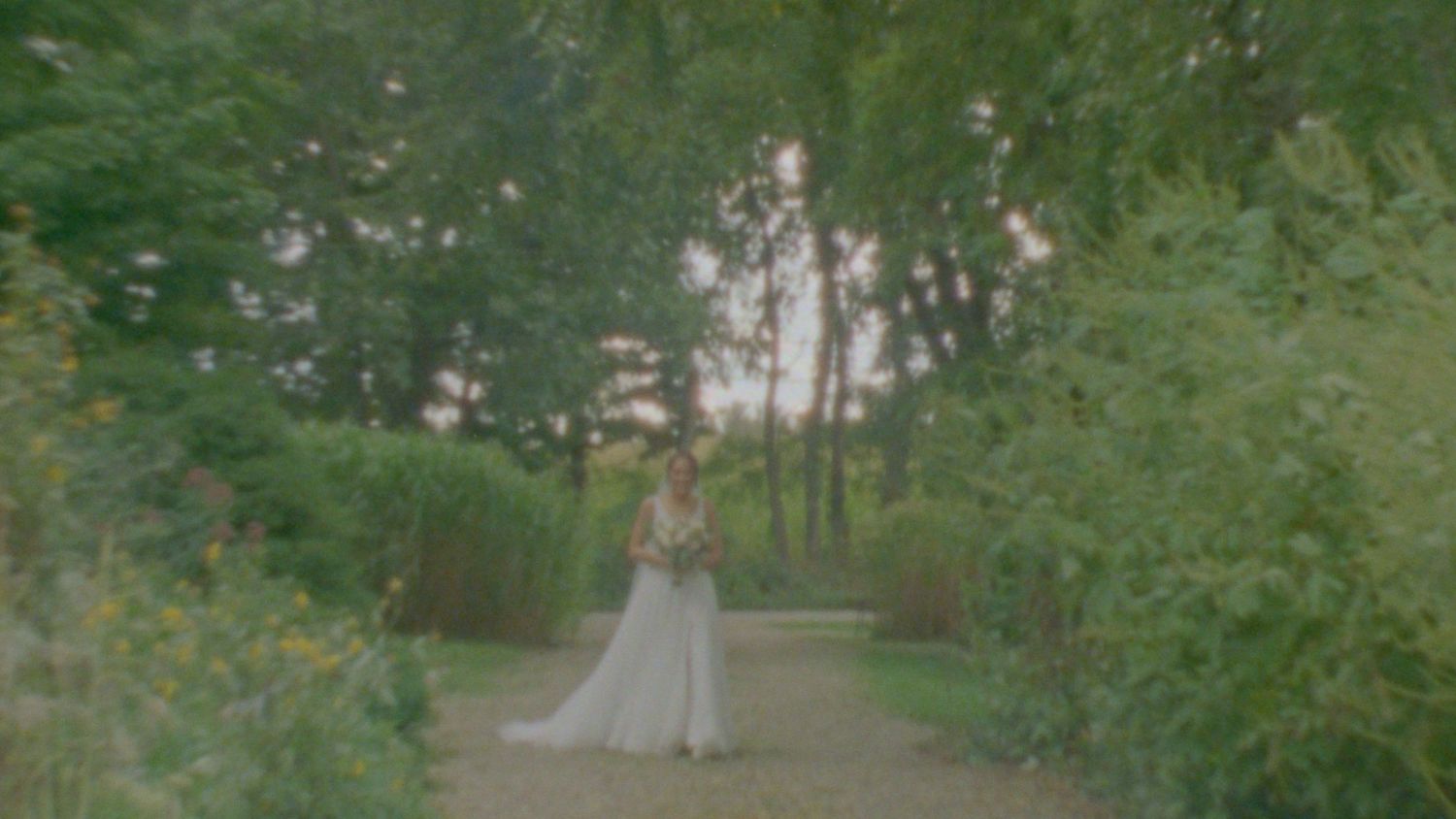 A bride in a white gown poses in a lush garden pathway surrounded by green trees and natural light.