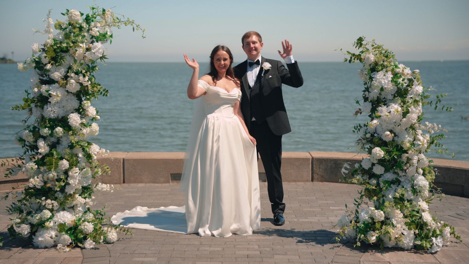 Bride and groom waving joyfully under floral arch with ocean backdrop on their wedding day.