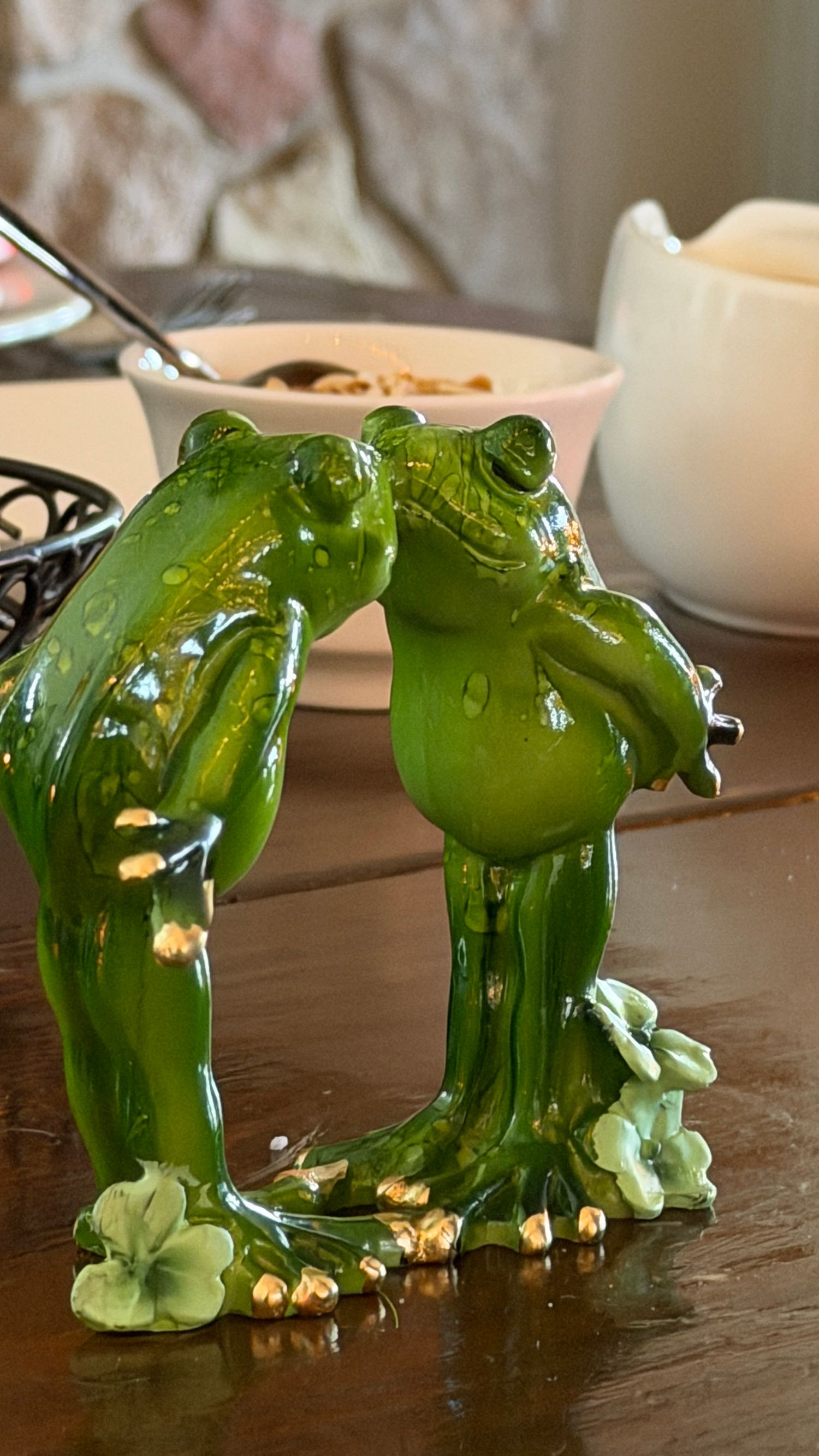 Two glossy green ceramic kissing frog figurines on a wooden table with a bowl in the background.