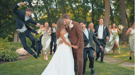 Joyful wedding party celebrates as bride and groom kiss outdoors, with guests jumping and cheering on lush green lawn.