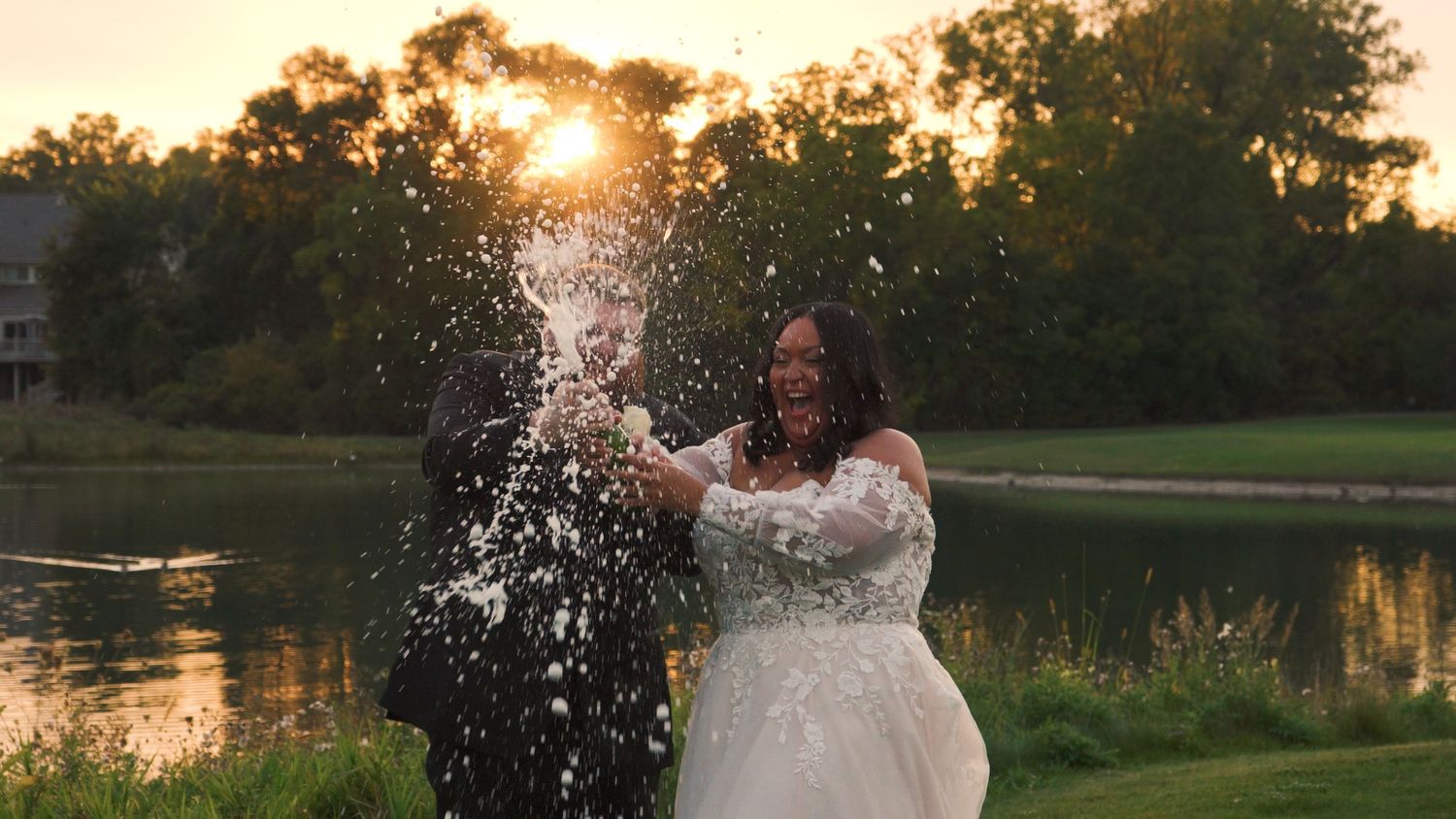 A joyful bride in a white lace dress splashes water by a lake at sunset creating a magical golden moment.