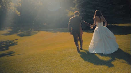 Bride and groom walking hand in hand across sunlit green lawn surrounded by trees on their wedding day.