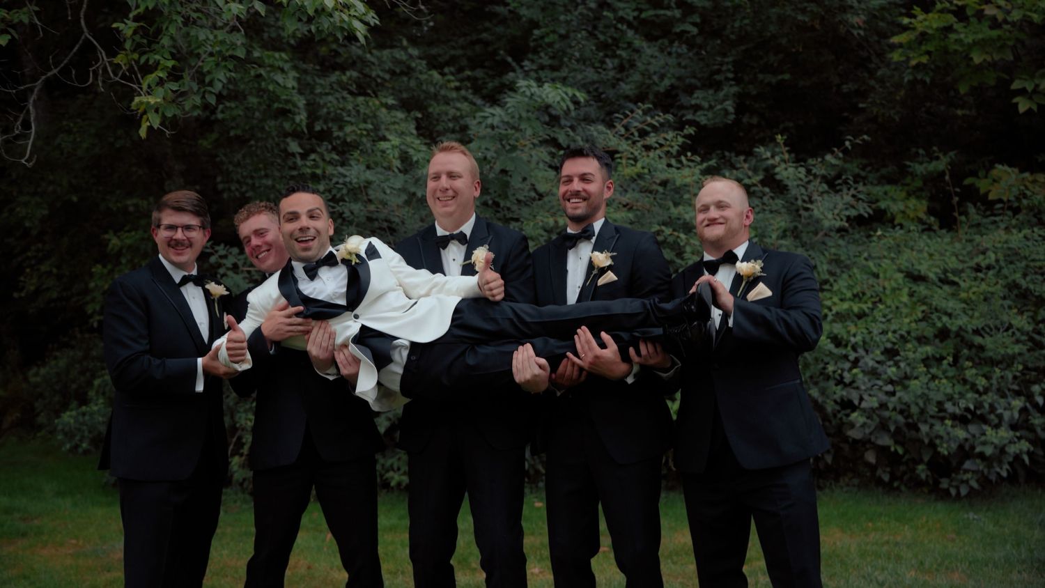 Groomsmen in black tuxedos lifting the groom horizontally outdoors, celebrating with smiles in front of lush green trees.