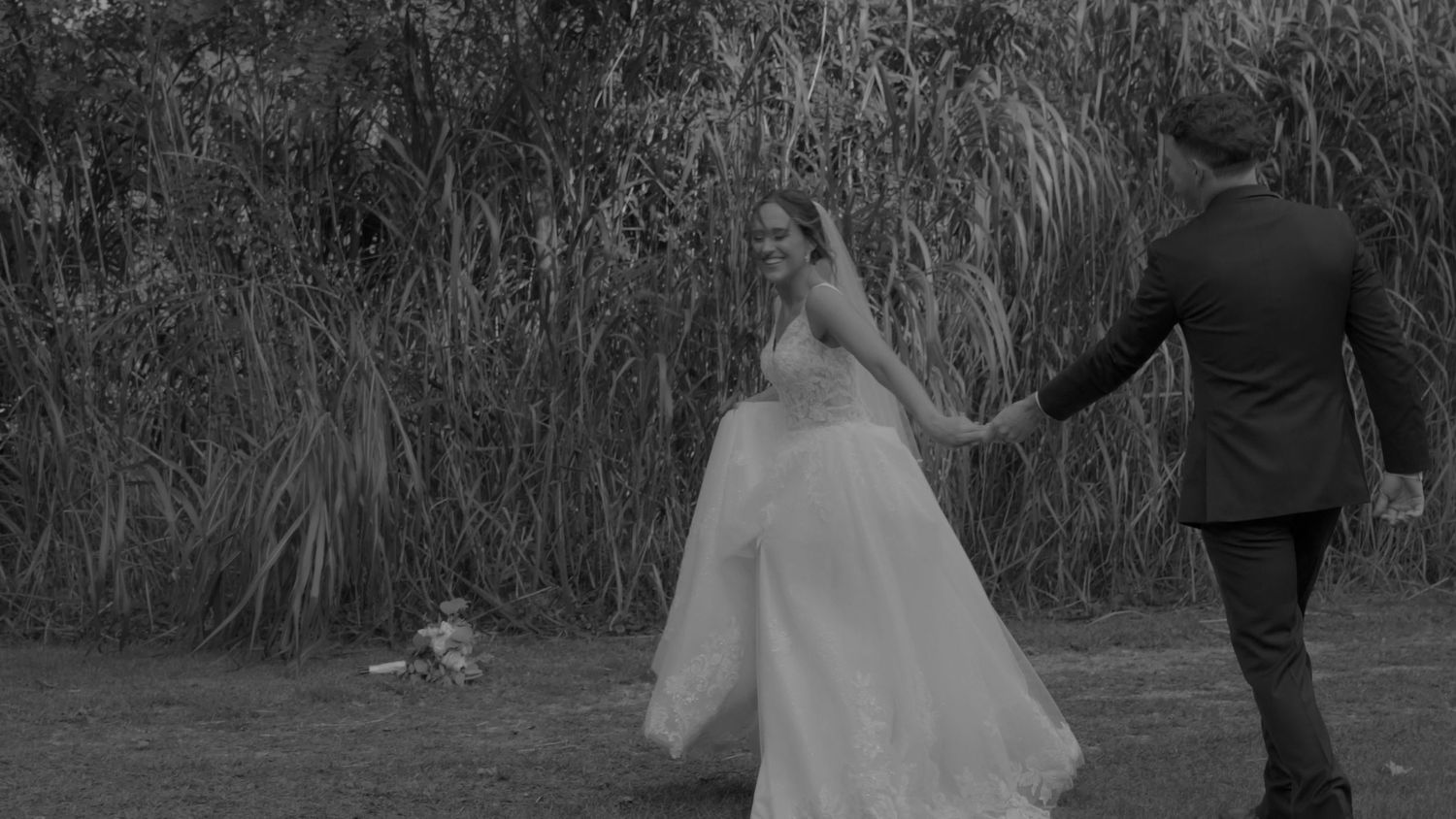A couple walks through tall grass holding hands in a romantic outdoor setting at sunset.